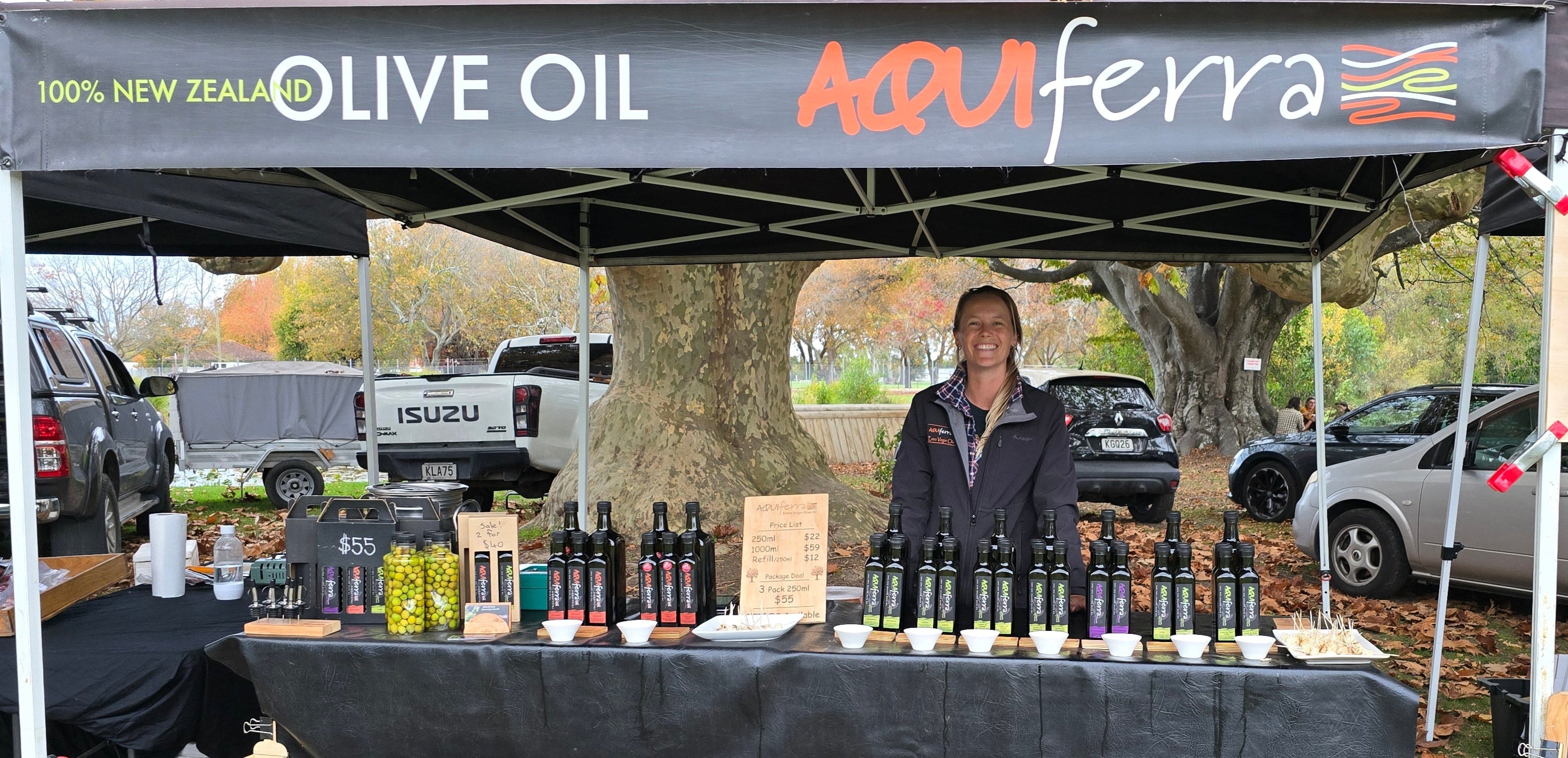 Person standing behind a table with olive oil bottles at an outdoor event, under a 'Aquiferra' olive oil tent.